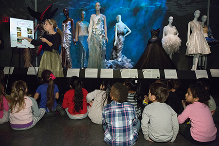 group of kids sitting on floor looking up at mannequins and teacher